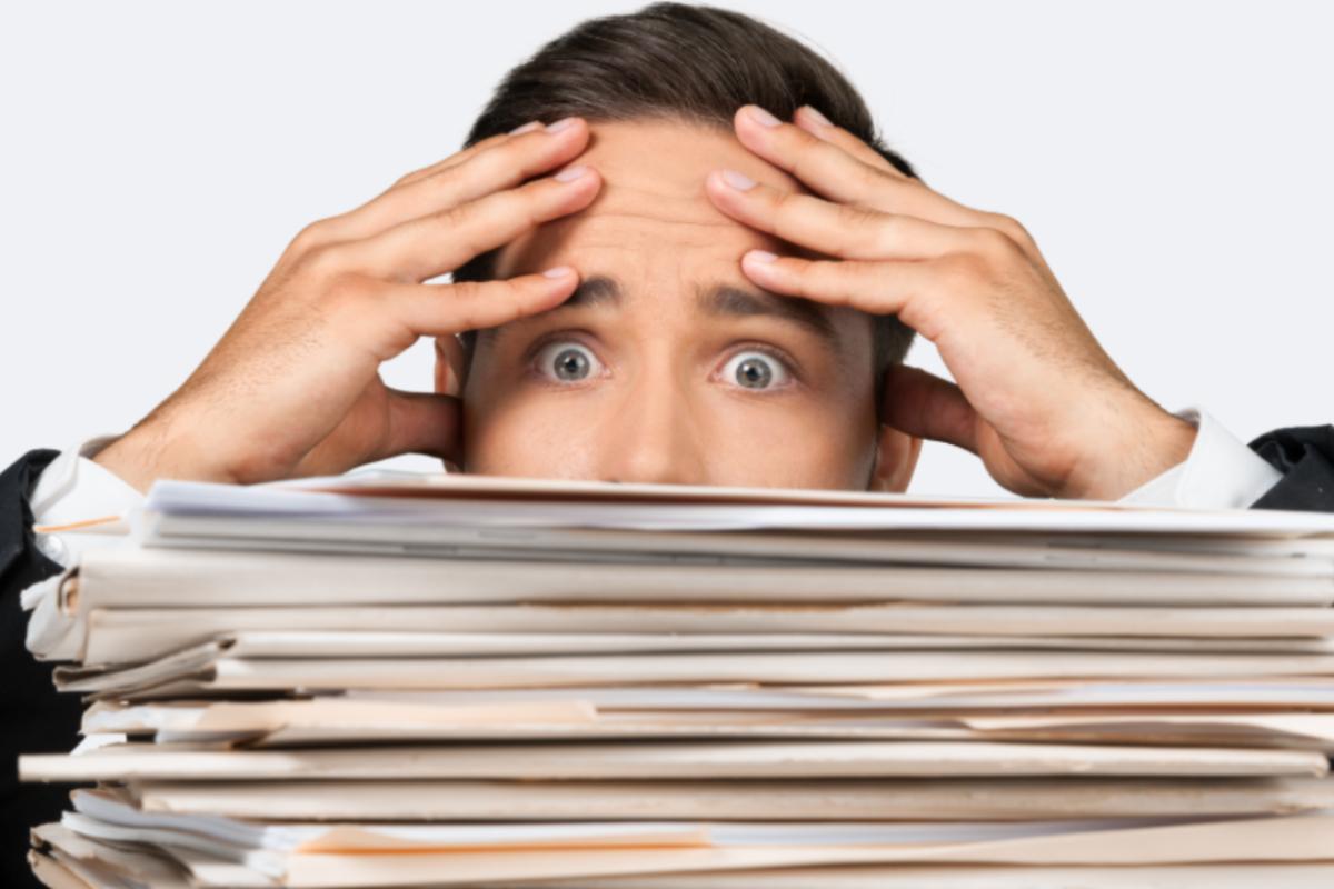 uomo preoccupato con le mani sulla fronte e il viso semi nascosto da fogli di lavoro